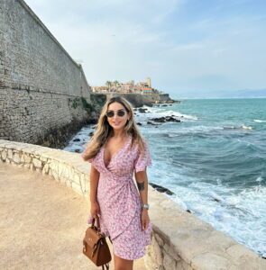 Women in pink floral dress enjoying stunning coastal view with historic castle and ocean waves in Antibes, France.