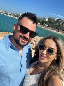 Beachside couple in sunglasses enjoying a sunny day in a coastal city with modern buildings in Cannes, France.