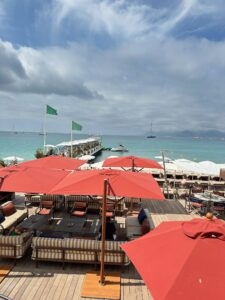 Colorful beachfront lounge with red umbrellas overlooking turquoise sea and sailing boats in Cannes, France.
