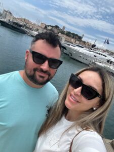 Smiling couple in sunglasses at marina in Cannes, France with yachts and historic buildings in the background.