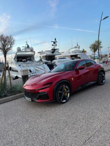 Luxury red Ferrari sports car at marina with yachts and boats docking, scenic waterfront view.