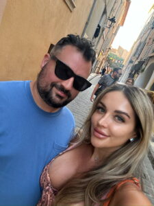 Smiling couple taking selfies in a charming European street with colorful buildings and cobblestone pavement.