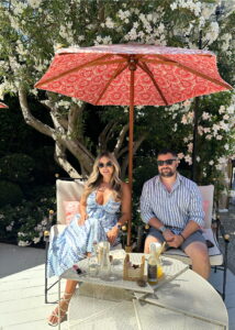 Relaxing outdoor patio scene with a woman and man enjoying drinks under a red umbrella amidst lush greenery Dior Restaurant Saint Tropez, France.