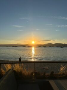 Sunset over a calm harbor with boats, mountains in the background, and person relaxing on the beach in Saint Tropez, France.