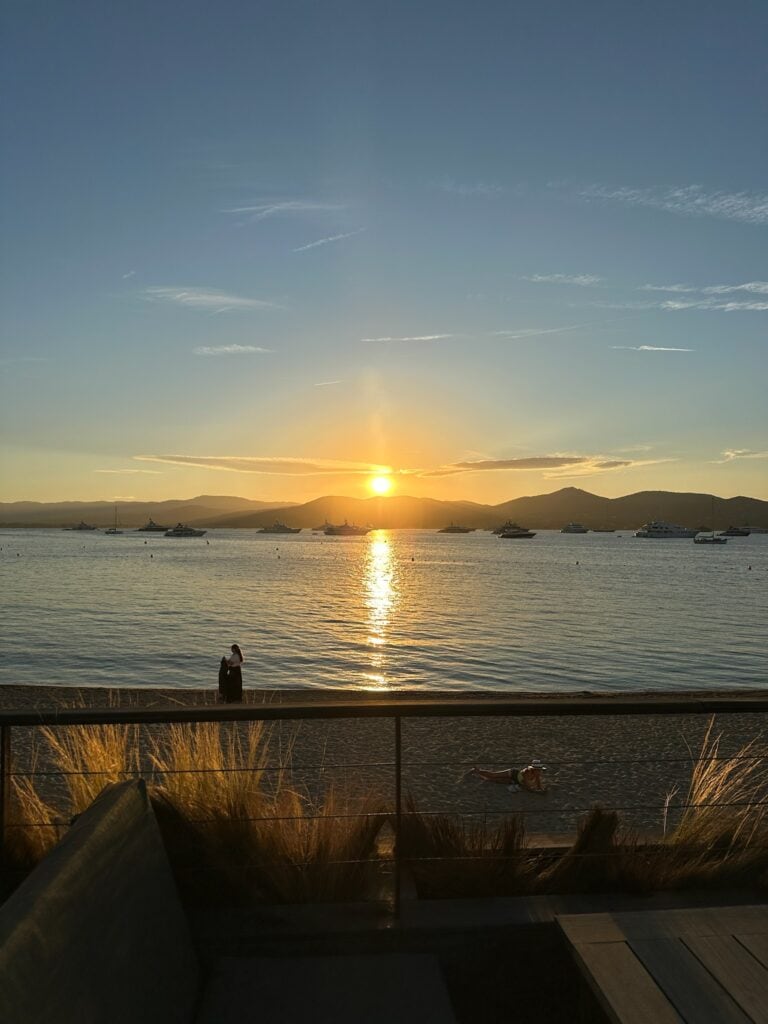 Sunset over a calm harbor with boats, mountains in the background, and person relaxing on the beach in Saint Tropez, France.