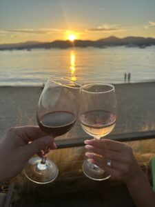 Romantic sunset at the beach with wine glasses clinking, showcasing scenic ocean and mountain views.