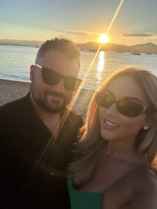 Silhouette of a couple enjoying sunset at the beach with mountains and boats in background.