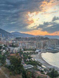 Panoramic view of Monaco coastline and cityscape at sunset, with mountains in the background and lush greenery.