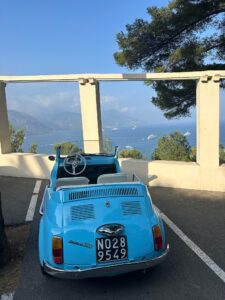 Vintage blue Fiat 500 convertible with scenic coastal view, boats sailing, and lush trees in the background.