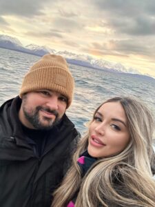 Bearded man and blonde woman taking a selfie on a boat in Norway with snowy mountains in the background.