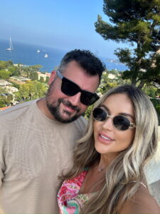 Man and woman in sunglasses enjoying scenic coastal view at sunset, smiling happily.