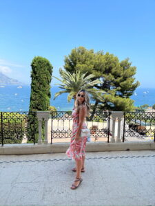 Stunning woman in a floral dress enjoying a scenic coastal view with palm trees and blue sea.